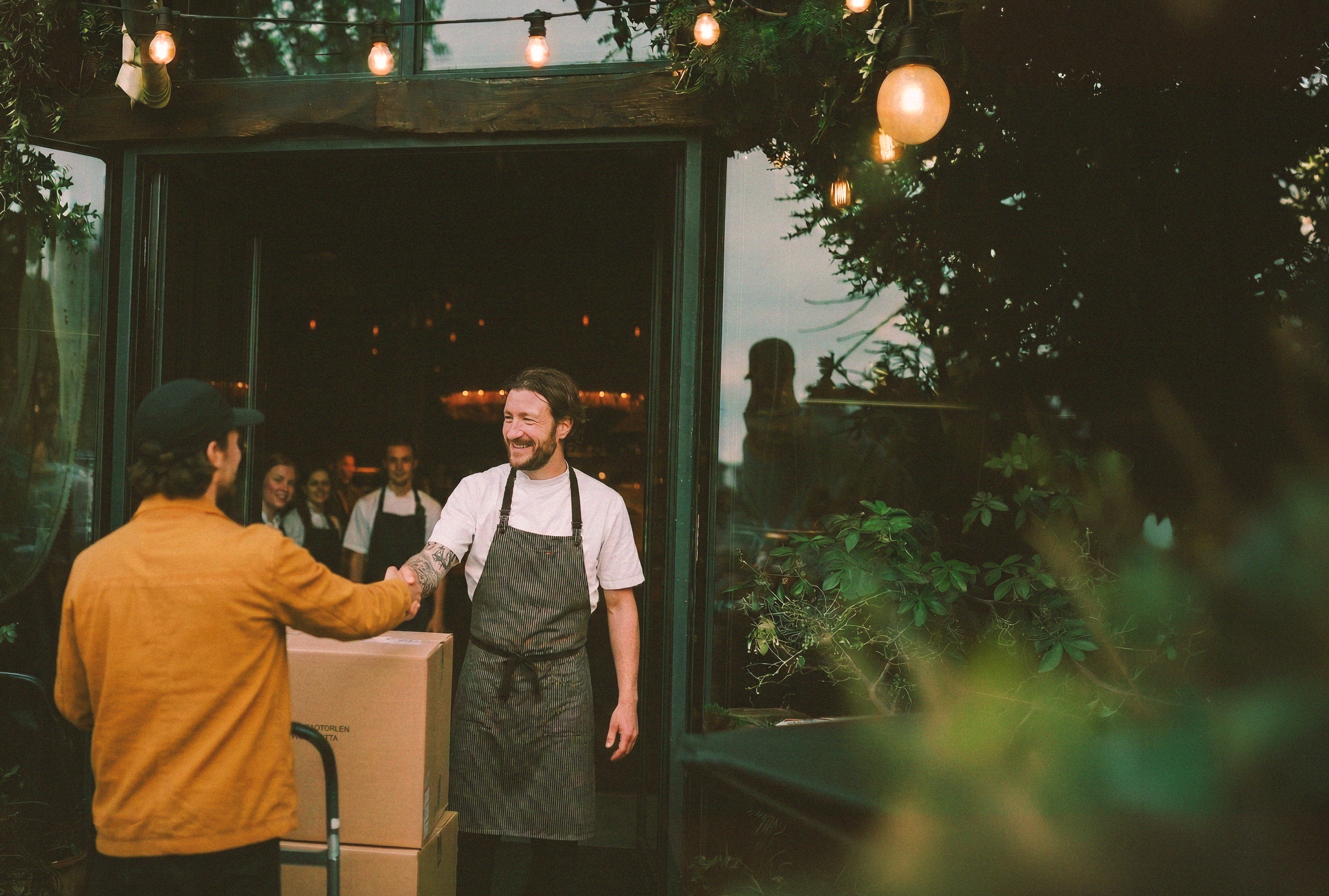 Man in apron shaking hands with another person outside a food truck with greenery in the background
