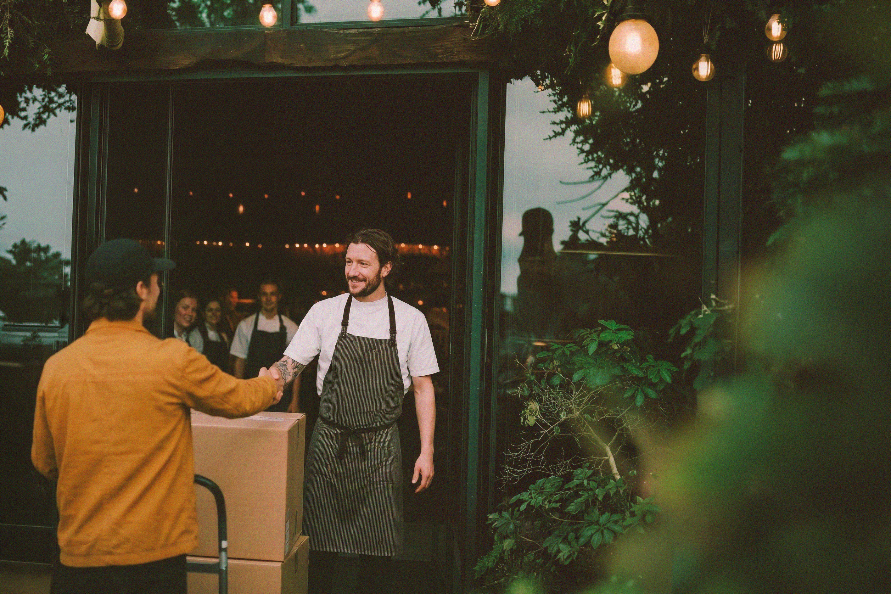 Two men shaking hands in front of a restaurant string lights and greenery.